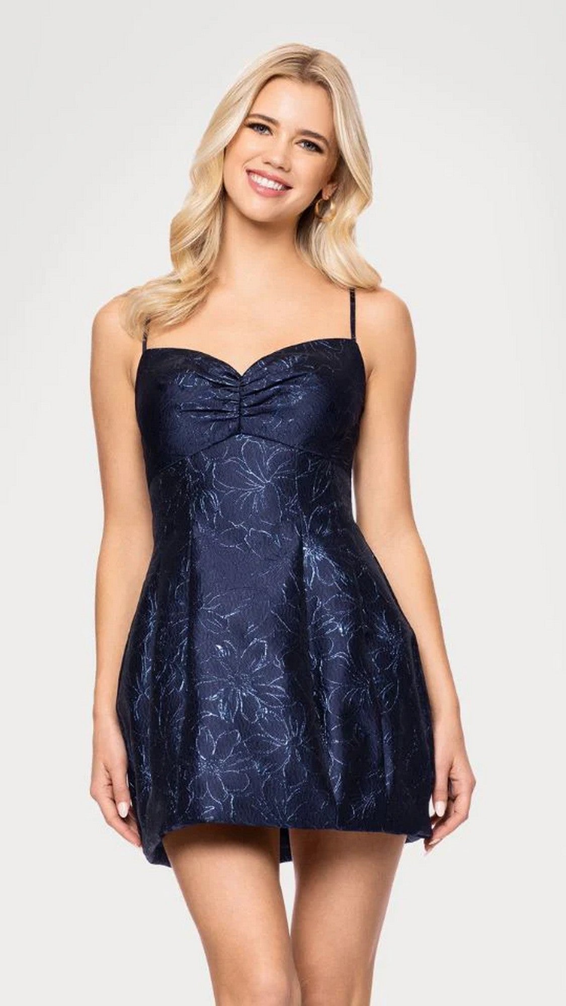 A woman smiles in the navy Short Prom Dress: Blondie Nites 5711BN, featuring thin straps, a sweetheart neckline, and a subtle metallic jacquard floral pattern.